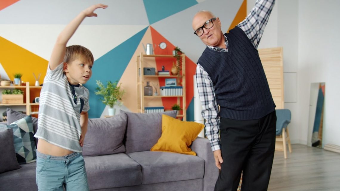 Retired grandfather and cute child do physical exercises together in house working out indoors caring for health. Healthy lifestyle and activity concept.